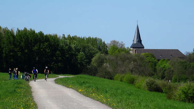 Mehrere Menschen sind mit Rädern unterwegs auf einem Weg, der zu einer Kirche im Grünen führt