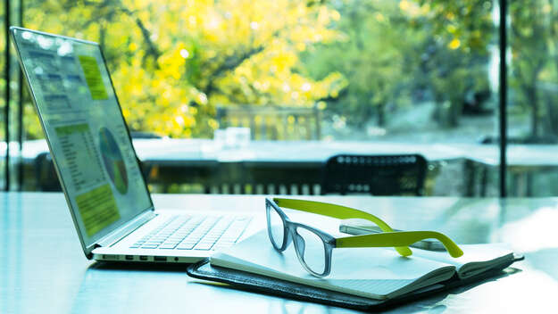 Aufgeklapptes Laptop und Brille vor einem fenster mit Blick ins Grüne