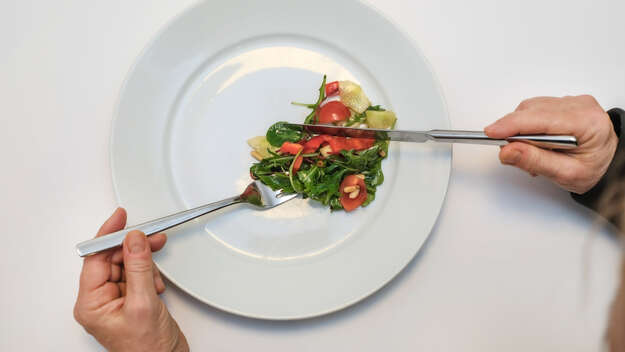 Zwei Hände mit Messer und Gabel über einem Teller mit etwas Salat
