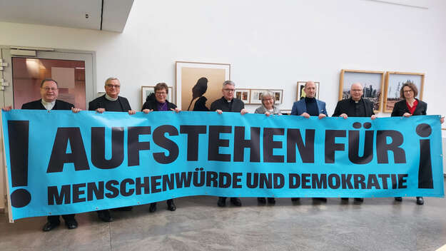 Vertreterinnen und Vertreter verschiedener Kirchen halten ein Banner hoch mit der Aufschrift 'Aufstehen für Menschenwürde und Demokratie' steht
