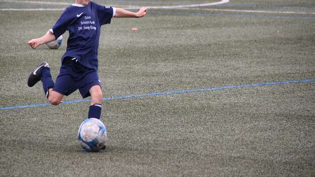 Junge beim Fußballspielen, der gerade im Lauf gegen den Fußball treten will. Der junge trägt ein blaues Trikot und eine blaue Sporthose.