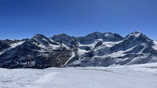 Berge und Skipiste in Samedan, Schweiz. Vorne im Bild die Skipiste, dahinter Berge und darüber ein blauer Himmel.