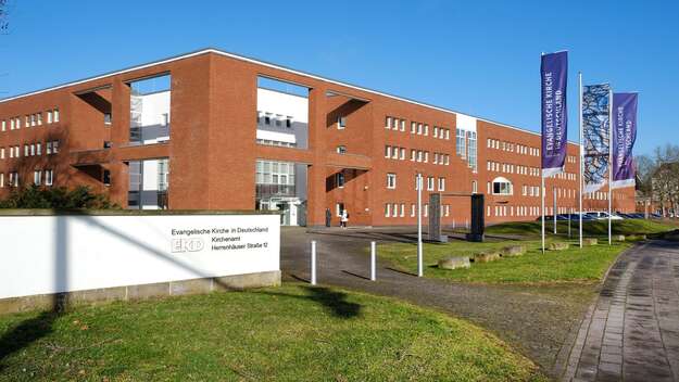 Zu sehen ist das Kirchenamt der EKD in Hannover. Ein braunes rechteckiges Gebäude. Davor wehen die lila Fahnen der EKD