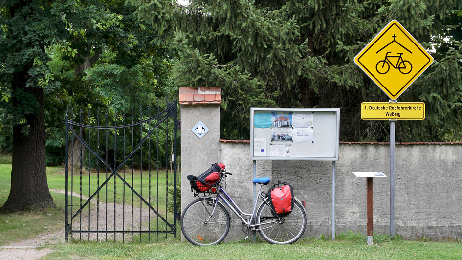 Bepacktes Fahrrad lehnt an der Mauer eines Kirchhofs neben dem Hinweisschild mit der Aufschrift '1. Deutsche Radfahrerkirche Wessnig' am Elbe-Radweg (Sachsen)