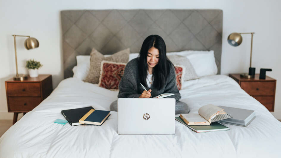 Eine junge Frau sitzt auf dem Bett und arbeitet am Laptop