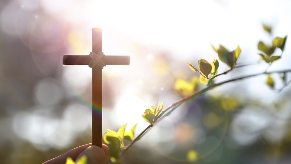 Eine Hand hält ein kleines Holzkreuz, das von warmem Sonnenlicht umgeben ist. Das Licht fällt weich durch den Hintergrund und erzeugt eine ruhige, hoffnungsvolle Stimmung. Neben dem Kreuz sind frische grüne Blätter zu sehen, die für neues Leben und Aufbru