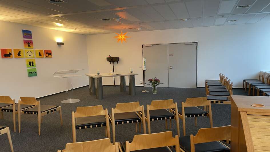 Zu sehen ist der Kirchenraum der evangelischen Kirche am Frankfurter Flughafen. Stuehle sind gestellt. Ein Altar ist zu sehen sowie ein buntes Kreuz an der linken, weissen Wand.