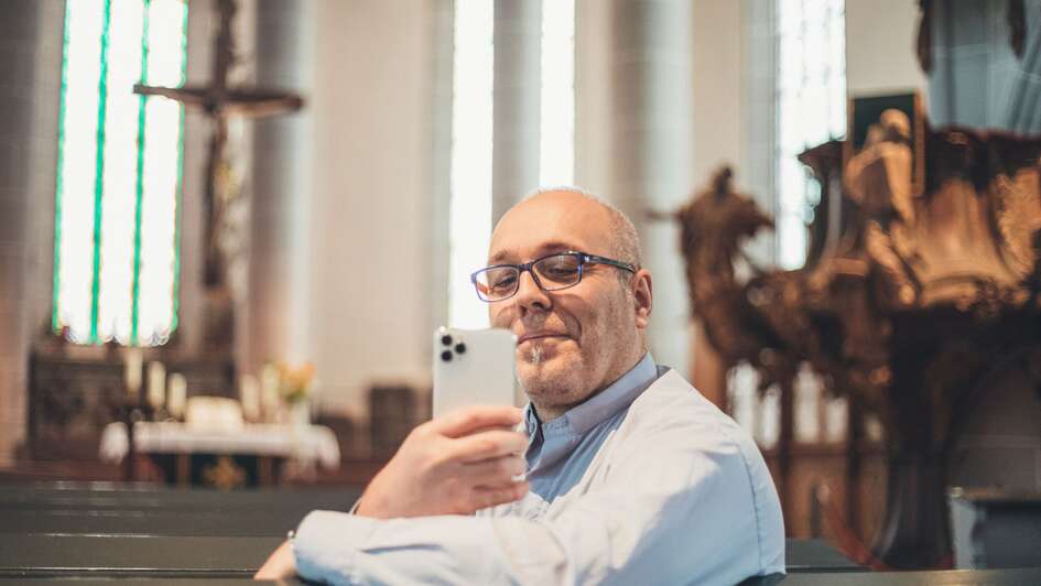 Zu sehen ist der Netzpfarrer Lutz Neumeier. Er sitzt in einer Kirche. In der Hand haelt er sein weisses Smartphone