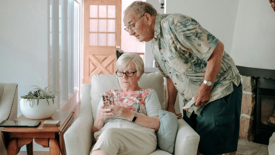 Älteres Paar in einem Wohnzimmer: Eine Frau sitzt auf einem Sessel und blickt konzentriert auf ihr Smartphone, während ein Mann sich über sie beugt und mit Interesse auf den Bildschirm schaut. Neben ihr liegt ein Tablet auf einem Beistelltisch.