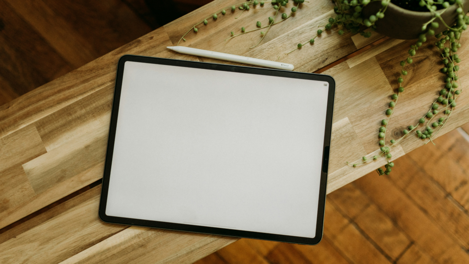 Tablet mit weißem Bildschirm und Stylus auf einem Holztisch neben einer hängenden Grünpflanze – aus der Vogelperspektive fotografiert.