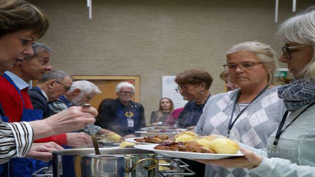 Ehrenamtliche Maenner und Frauen stehen entlang einer Tischreihe mit Catering Boxen. Sie legen Braten und Klösse auf die Teller